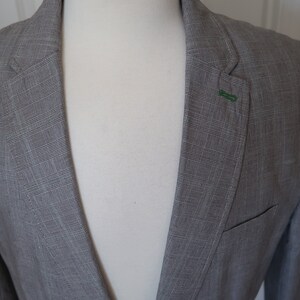 May include: A gray blazer with a subtle checkered pattern. The blazer has a single button closure and a green lapel pin.