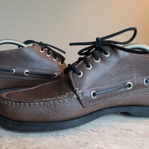 May include: A pair of brown leather boat shoes with black laces. The shoes have a moc toe design and a rubber sole.