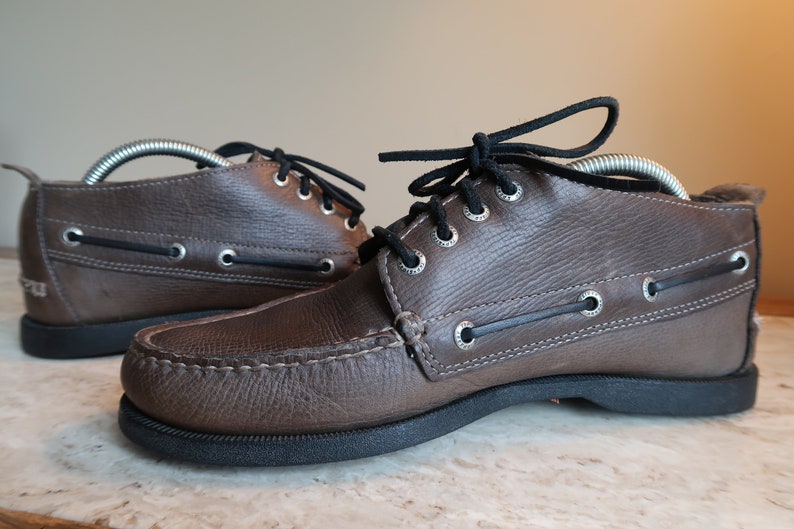 May include: A pair of brown leather boat shoes with black laces. The shoes have a moc toe design and a rubber sole.