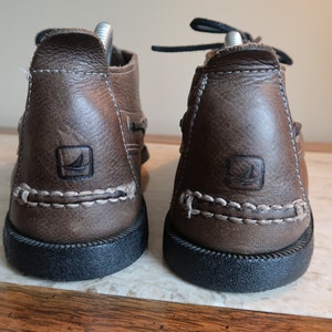 May include: A pair of brown leather boat shoes with a white stitching detail and a black rubber sole. The shoes have a logo on the side.