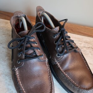 May include: A pair of brown leather boat shoes with black laces. The shoes have a stitched seam around the toe and a rubber sole.