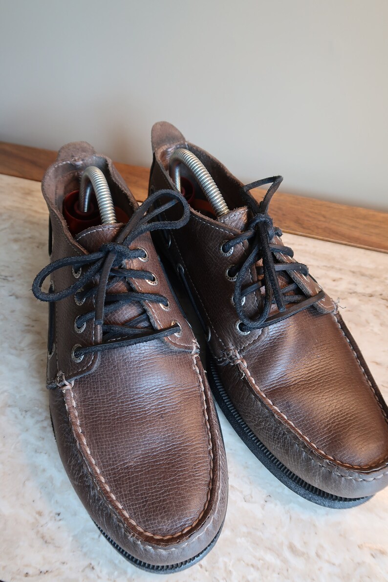 May include: A pair of brown leather boat shoes with black laces. The shoes have a stitched seam around the toe and a rubber sole.