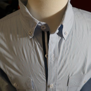 May include: A blue and white striped long-sleeve shirt with a dark blue and white trim down the front. The shirt has a button-down collar and two buttoned pockets.
