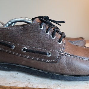 May include: Brown leather boat shoes with laces and a white stitching detail. The shoes have a black rubber sole and the text "Topsiders" is visible on the sole.