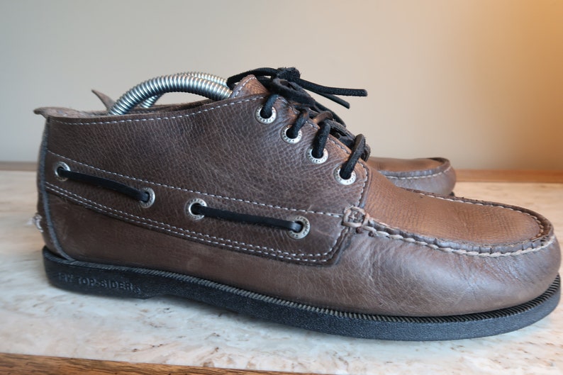 May include: Brown leather boat shoes with laces and a white stitching detail. The shoes have a black rubber sole and the text "Topsiders" is visible on the sole.