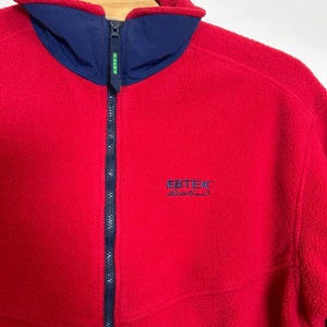 May include: A red fleece jacket with a navy blue collar and zipper. The jacket has the brand name "EBTEK" embroidered in navy blue on the chest. The jacket is a full-zip style.