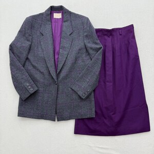 Può includere: Un blazer grigio con motivo a quadri viola e una gonna viola abbinata. Il blazer ha una fodera viola e chiusure con bottoni. La gonna è di colore viola unito.