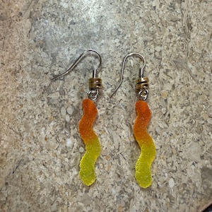 Könnte beinhalten: Ein Paar ausgefallene Ohrringe mit einem Gummibärchendesign. Die Ohrringe haben silberfarbene Haken und kleine goldfarbene Perlen. Die Gummibärchen sind ein Farbverlauf von Orange und Gelb.
