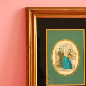 Puede incluir: Una imagen enmarcada con un marco dorado y un paspartú negro. La imagen es de un hombre y una mujer en un entorno de jardín. La mujer lleva un vestido azul y el hombre lleva un traje negro.