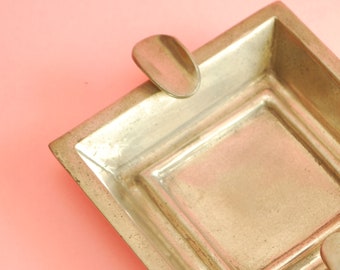 May include: A silver ashtray with a square shape and two small, rounded indentations on the sides. The ashtray has a raised, square design in the center.