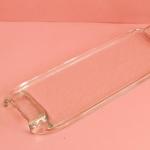 Könnte beinhalten: Ein rechteckiges Serviertablett aus klarem Glas mit abgerundeten Ecken und kleinen Griffen. Das transparente Tablett liegt auf einem rosa Hintergrund. Das Design ist schlicht und elegant, geeignet zum Servieren von Speisen oder zum Präsentieren von Gegenständen.