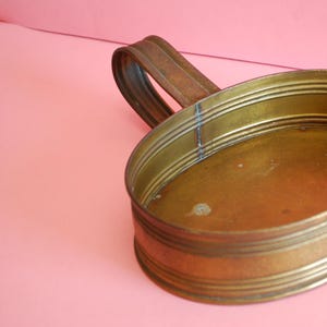 Puede incluir: Un plato ovalado de latón con un asa. El plato tiene una superficie lisa y brillante y está decorado con una serie de anillos elevados alrededor del borde.