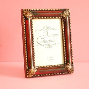 Peut inclure: Un cadre photo rouge et doré avec des détails ornés et le texte "Antique Collection" sur le devant. Le cadre mesure 6,35 x 8,89 cm.
