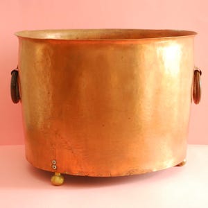 May include: A large copper planter with two black handles and three brass feet. The planter has a hammered finish and is oval in shape.