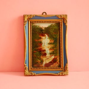 Może przedstawiać: Oprawiony obraz krajobrazu wodospadu kaskadowego przez las. Obraz jest w prostokątnej ramie z niebieską obwódką i złotymi akcentami. Grafika jest wyświetlana na różowym tle.