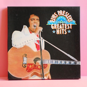 Peut inclure: Une pochette d'album en noir et blanc avec une photo d'Elvis Presley chantant dans un microphone tout en tenant une guitare. Le titre de l'album est "Elvis Presley's Greatest Hits" avec des étoiles autour des mots "Greatest Hits".