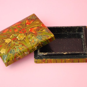 Może przedstawiać: Otwarte drewniane pudełko z kwiatowym wzorem na pokrywie. Pudełko jest pomalowane w odcieniach zieleni, żółci, czerwieni i czerni. Wnętrze pudełka jest czarne.