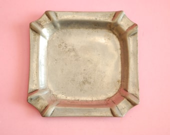 May include: A silver metal ashtray with a square shape and rounded edges. The ashtray has a slightly worn surface.