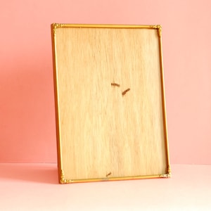 May include: A rectangular gold-colored picture frame with ornate corner detailing. The frame has a light-colored wooden backing. The frame is leaning against a pink background.