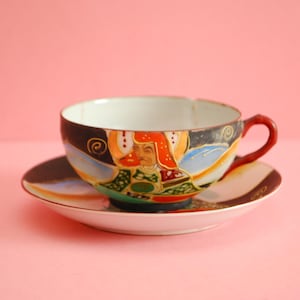 Op de afbeelding: Een vintage theekopje en schotelset met een kleurrijk handgeschilderd ontwerp met een persoon in traditionele Japanse kleding. Het kopje en de schotel zijn beide wit met een zwarte rand.