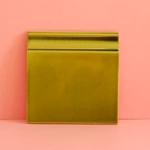Può includere: Una piastrella quadrata in ceramica verde oliva con finitura lucida. La piastrella ha un bordo superiore arrotondato e rialzato. Lo sfondo è di colore rosa chiaro uniforme. Questa piastrella è probabilmente per l'arredamento della casa o la costruzione.