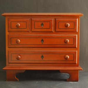 Peut inclure: Une commode en bois avec quatre tiroirs. Les tiroirs sont ornés de bordures noires et d'un motif en forme de diamant au centre. La commode est d'une couleur brun rougeâtre riche.