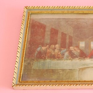 Op de afbeelding: Een ingelijste print van Het Laatste Avondmaal, een beroemd schilderij uit de Renaissance van Leonardo da Vinci. De print is in een gouden en witte lijst met een touwachtig ontwerp.