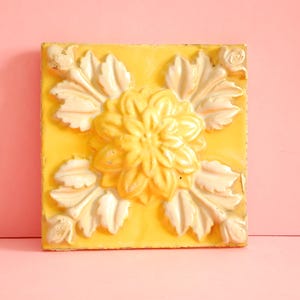 Può includere: Piastrella decorativa quadrata gialla con un motivo floreale in rilievo. Il fiore centrale è circondato da foglie stilizzate di un colore bianco sporco più chiaro. La piastrella è posta su uno sfondo rosa.
