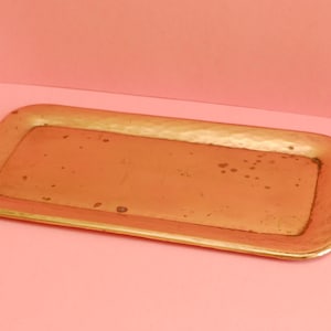 Puede incluir: Una bandeja rectangular de metal de color dorado con un acabado martillado. La bandeja es rectangular y tiene un borde ligeramente elevado.