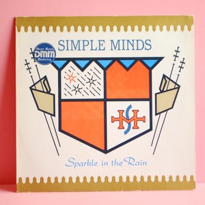 Peut inclure: Une pochette d'album vinyle blanche avec une bordure dorée. La pochette présente un blason avec un écu bleu et rouge, une bannière bleue et blanche et un fond blanc avec des étoiles noires. Le texte "SIMPLE MINDS" est imprimé en lettres noires au-dessus du blason. Le texte "Sparkle in the Rain" est imprimé en lettres noires en dessous du blason. La pochette d'album présente également un petit autocollant bleu et blanc avec le texte "Direct Metal Mastering DMM" imprimé en lettres blanches.