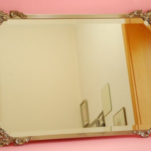 May include: A large, ornate, silver-colored mirror with a decorative, floral-patterned frame. The mirror is reflecting a room with several framed pictures.