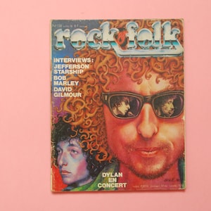 Puede incluir: Portada de revista vintage "rock folk" con retratos de músicos. La portada incluye el título de la revista, listas de entrevistas y la frase "Dylan en Concert". La revista está sobre un fondo rosa.