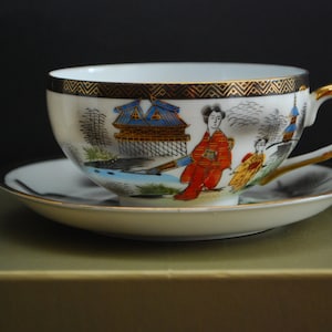 Op de afbeelding: Een wit theekopje en schotel met een gouden rand en een Japans geïnspireerd ontwerp met een vrouw in een rode kimono en een blauw-witte pagode.