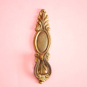 Puede incluir: Un tirador de cajón de metal dorado con un diseño ornamentado. El tirador tiene una manija en forma de anillo y una forma ovalada decorativa en la parte superior.