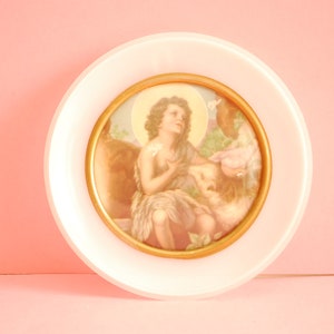 Puede incluir: Un plato de cerámica blanco con una imagen enmarcada en oro de una persona joven con cabello largo, vestida con una túnica marrón, sosteniendo un cordero. La imagen está sobre un fondo marrón claro.
