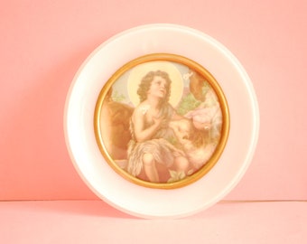 May include: A white ceramic plate with a gold-framed picture of a young person with long hair, wearing a brown robe, holding a lamb. The picture is set against a light brown background.