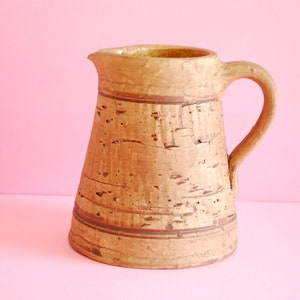 May include: A brown ceramic pitcher with a textured surface and a brown handle. The pitcher has a wide base and a narrow neck.