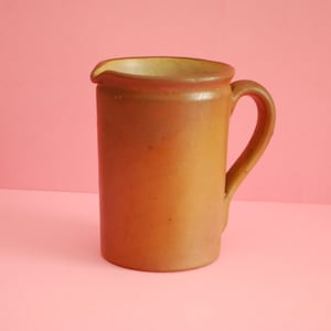 Könnte beinhalten: Ein brauner Keramik-Krug mit Henkel. Der Krug steht auf einem rosa Hintergrund.