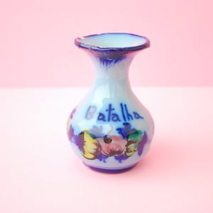 Puede incluir: Un pequeño jarrón de cerámica azul claro con un borde acampanado y un cuerpo redondeado. El jarrón está decorado con diseños florales coloridos y la palabra "Batalha" en escritura azul. El jarrón tiene un borde y una base azul oscuro.