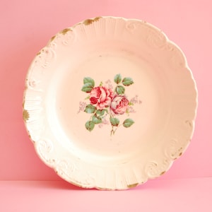 May include: A white porcelain plate with a floral design featuring pink roses and green leaves. The plate has a scalloped edge and a raised rim.