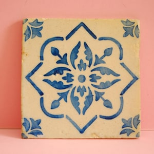 Puede incluir: Un azulejo de cerámica blanco con un patrón floral azul y blanco. El azulejo tiene un diseño geométrico con una flor central y cuatro hojas que la rodean.