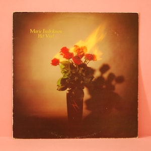 Puede incluir: Portada de álbum de vinilo vintage con un jarrón de rosas rojas con llamas. El álbum se titula "Het Vind" de Marie Fredriksson. El fondo es un degradado marrón y amarillo.