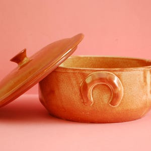 Peut inclure: Un plat à casserole en céramique orange ouvert avec un couvercle assorti. Le plat a une forme rectangulaire arrondie avec une poignée de chaque côté. Le couvercle est légèrement entrouvert, révélant l'intérieur.