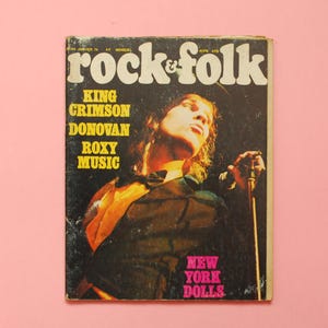 Puede incluir: Portada de la revista vintage "rock & folk" con un músico en el escenario. Se ven el título y nombres de bandas como King Crimson y Roxy Music. El fondo es rosa.