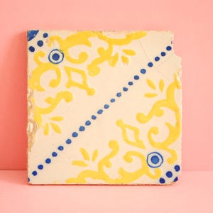 Puede incluir: Un azulejo de cerámica blanco con un patrón floral amarillo y azul. El azulejo tiene un aspecto desgastado, con parte de la pintura descascarada.