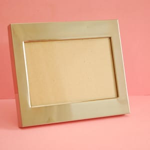 Può includere: Una cornice rettangolare color argento con un interno color beige chiaro. La cornice è leggermente angolata, mostrando le sue linee pulite e il design minimalista. Lo sfondo è rosa.