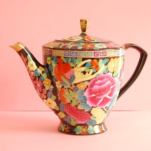 Puede incluir: Una tetera negra decorativa con tapa, asa y boquilla. La tetera está adornada con coloridos diseños florales y frutales, incluyendo una gran flor rosa. La boquilla y el asa de la tapa tienen detalles dorados.