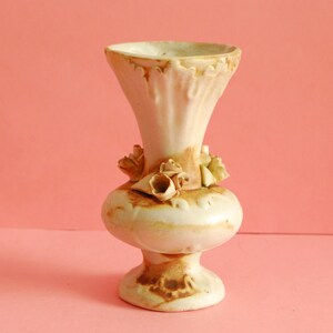 May include: A small, white ceramic vase with a brown glaze and floral accents. The vase has a wide mouth and a narrow base.