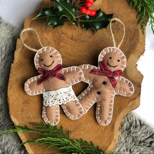 May include: Two felt gingerbread people ornaments with red and white plaid bows. The gingerbread person on the left is wearing a white lace trim. Both ornaments have black button eyes and smiles.