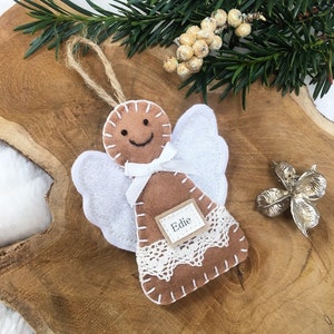 May include: A brown felt gingerbread angel ornament with white felt wings and a white ribbon. The ornament has a brown felt tag with the name "Edie" written on it.